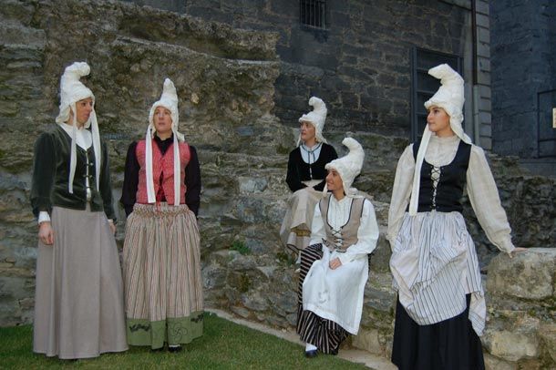 Cinco mujeres con trajes vascos antiguos y altos tocados blancos de tela enrollada, como el gorro de Mari Domingi, junto a un muro de piedra