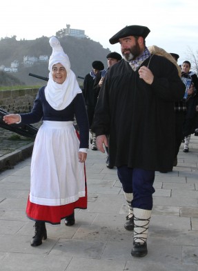 Mari Domingi, con tocado blanco y delantal, camina sonriendo junto a Olentzero, con txapela y chaqueta negra, en un desfile de Navidad